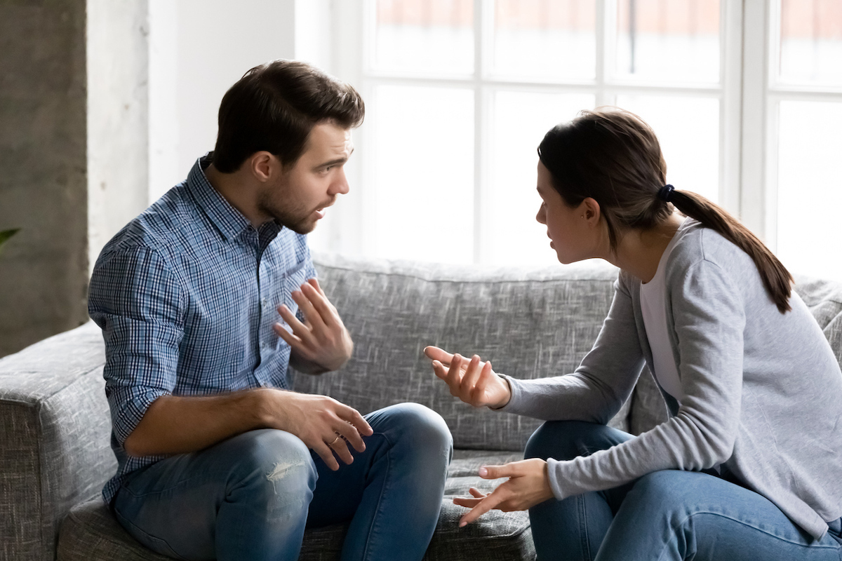 A young couple arguing, woman pointing blame at man, while sitting on their couch.