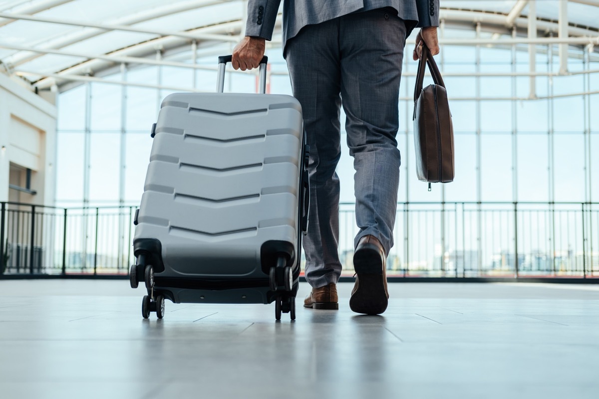 Low angle view of unrecognizable businessman in gray suit walking through airport terminal with briefcase and suitcase.