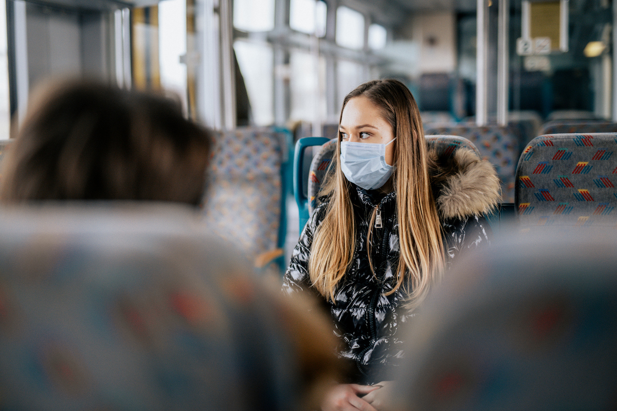 Young woman is wearing protective face mask while coronavirus is around. She is sitting on a bus or train. Photo is taken over the shoulder of another person. The woman in the main image is looking outside the window.