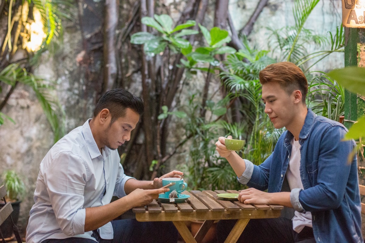 young man looking at friend using mobile phone in cafe. Males are having beverages at table. They are sitting together in coffee shop. (Young man looking at friend using mobile phone in cafe. Males are having beverages at table