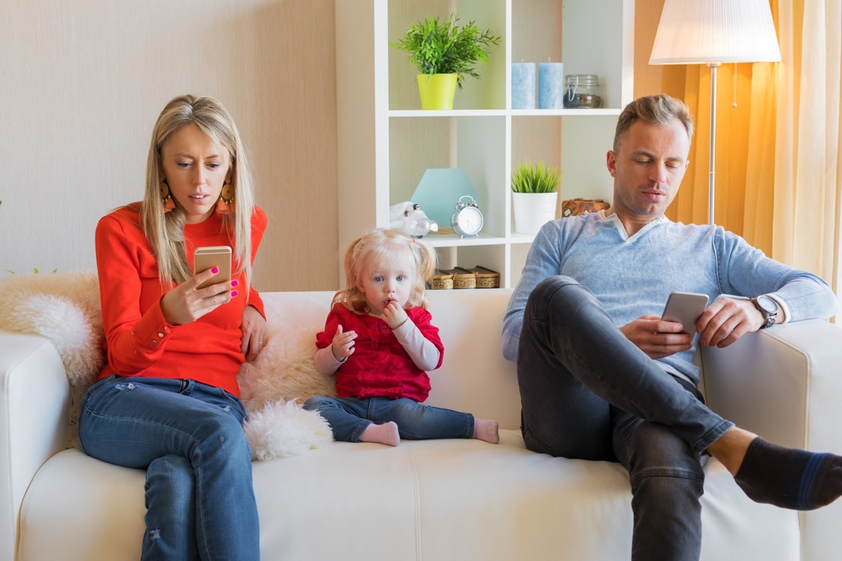 mom and dad sitting on a white couch playing with cell phones in front of their young daughter