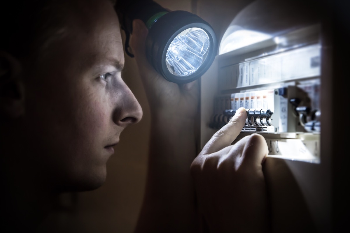 Horizontal composition photography of a young men, specialized technician or just home man, trying to restore power after electricity outage in the dark with his light. He is looking on the circuit breaker, and try to find the right button to operate. Picture shot in the dark, with a LED hand light, which gives a cold colorimetry image, and selective focus on the finger human hand and button of the circuit breaker (ON-OFF), fuse box switch. Electrical cabinet, open, with electrical circuit breaker. Side view in selective focus.