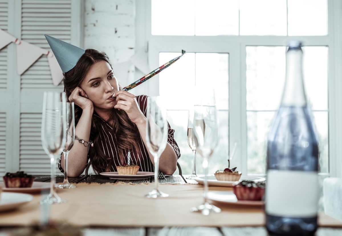 woman looking forlorn and sitting alone at a table wearing a party hat and blowing a noisemaker