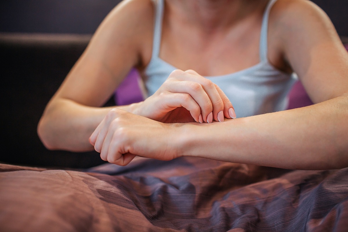 Cut view of young woman sitting on bed and touching her skin with one hand. She is in room alone. Her legs are covered with blanket. Woman has problem