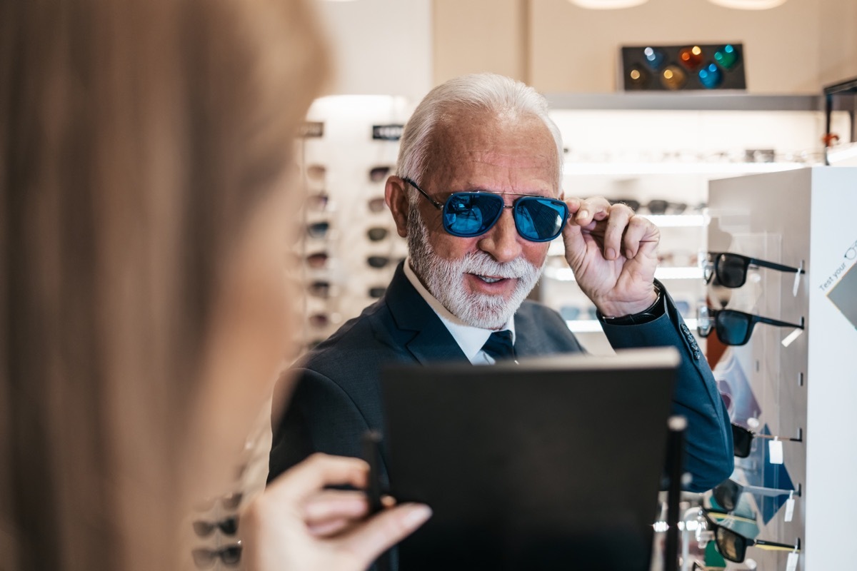 Elegant senior businessman choosing and buying sunglasses in optical store and young female seller helping him to make right decision.