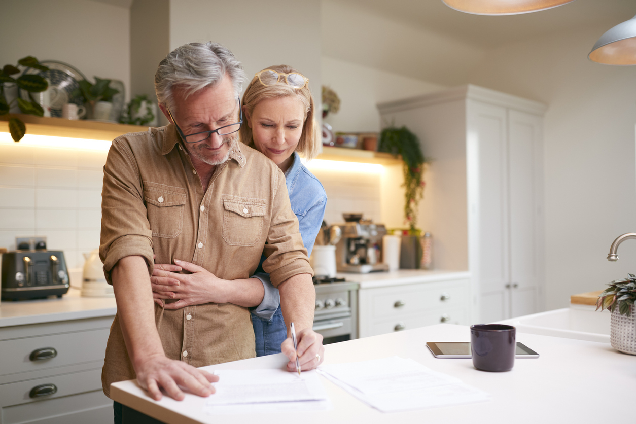 A middle-aged couple standing at their kitchen councer and looking over documents while hugging