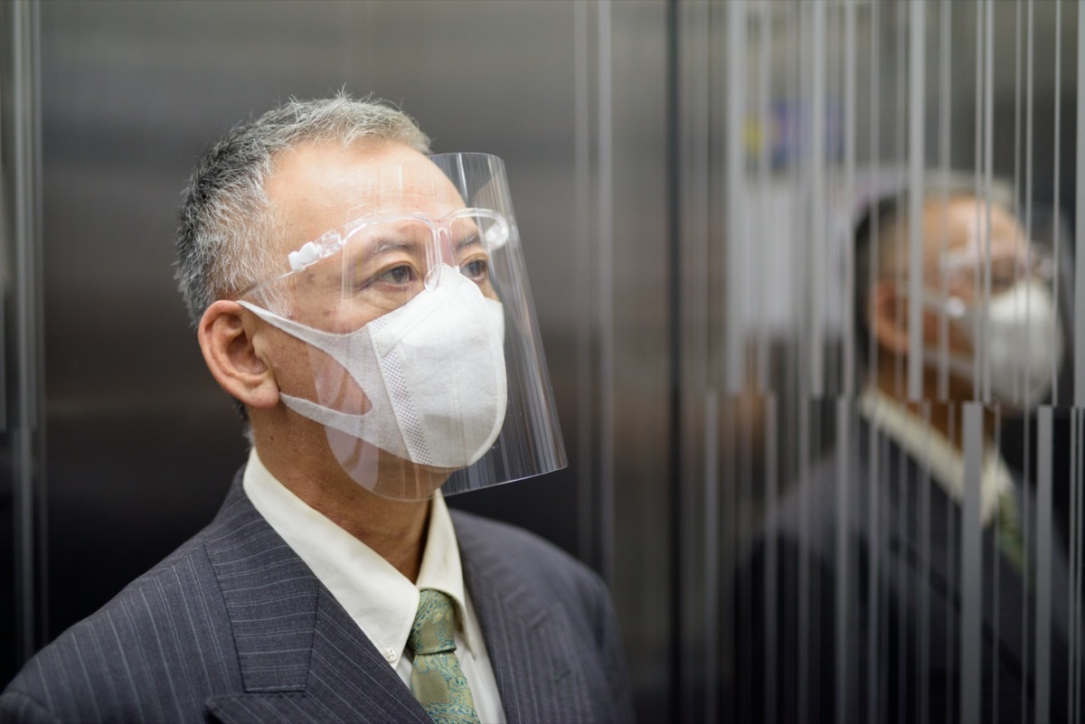 older asian businessman riding elevator while wearing face shield amid coronavirus