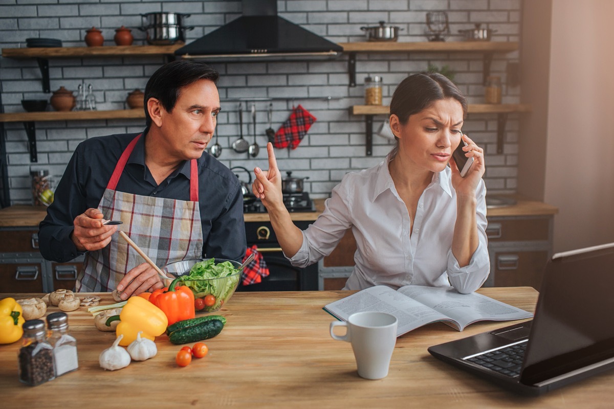 Latino man wearing apron interrupts wife on phone call in the kitchen, etiquette over 40