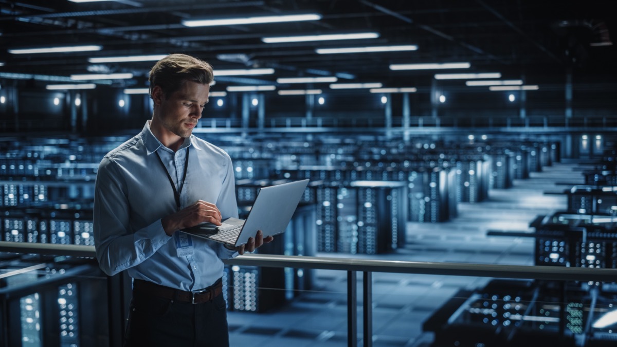 Portrait of IT Specialist Uses Laptop in Data Center. Server Farm Cloud Computing Facility with Male Maintenance Administrator Working. Cyber Security and Network Protection.