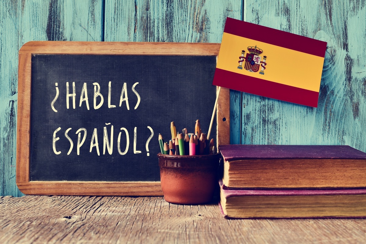 a chalkboard with the question hablas espanol? do you speak Spanish? written in Spanish, a pot with pencils and the flag of Spain, on a wooden desk