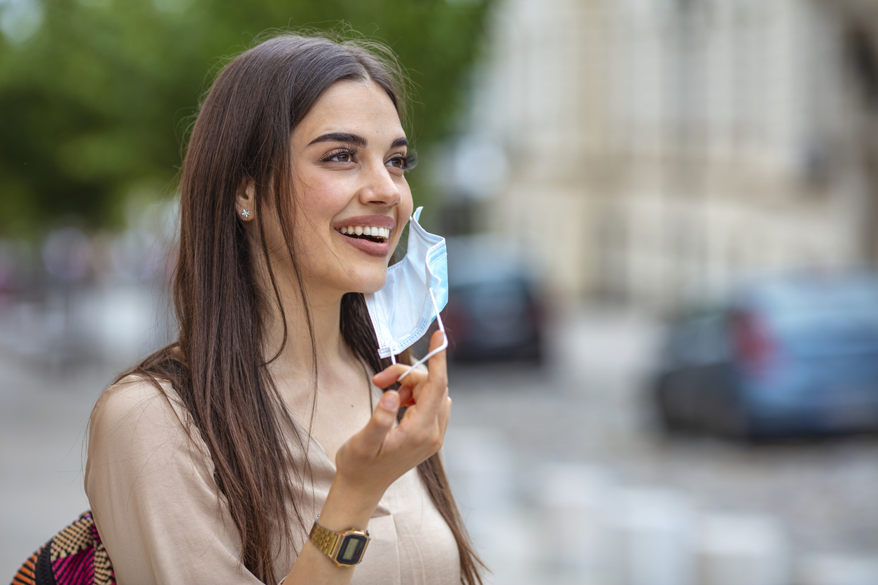 A young woman removing her face mask while standing outdoors.