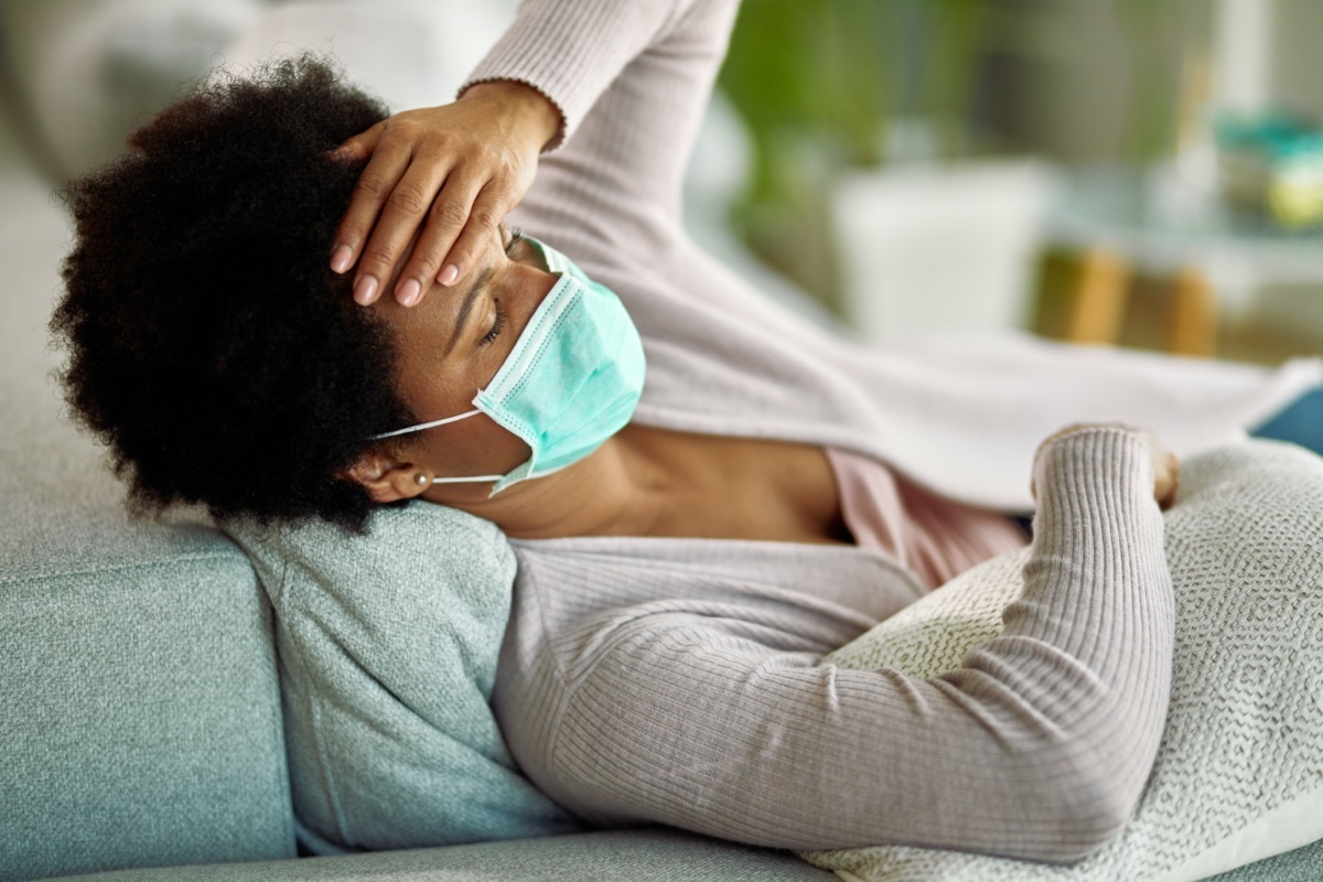 Tired black woman with face mask having a headache while lying down on sofa at home.