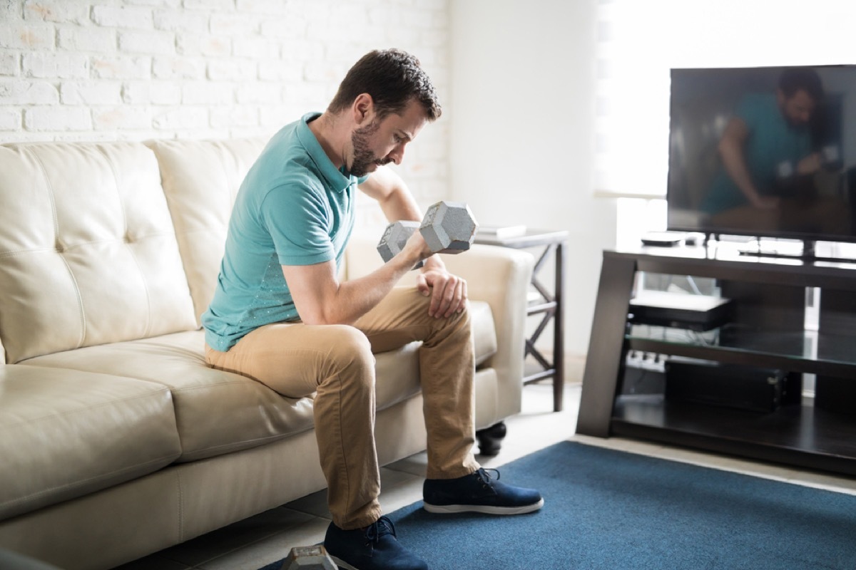 white man lifting weights while sitting on his couch in front of tv