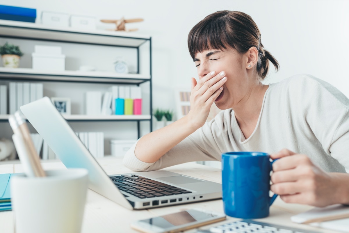 Tired sleepy woman yawning, working at office desk and holding a cup of coffee, overwork and sleep deprivation concept