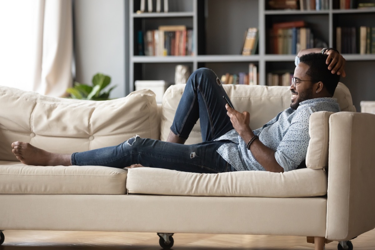 Full length relaxed young man in glasses lying on comfortable couch, enjoying spending weekend leisure time chatting in social network on phone with friends, watching funny videos.