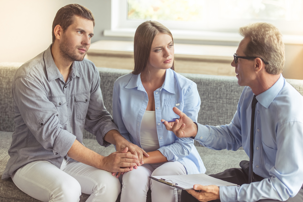 A young couple holding hands shares the couch with their therapist who's talking to them.