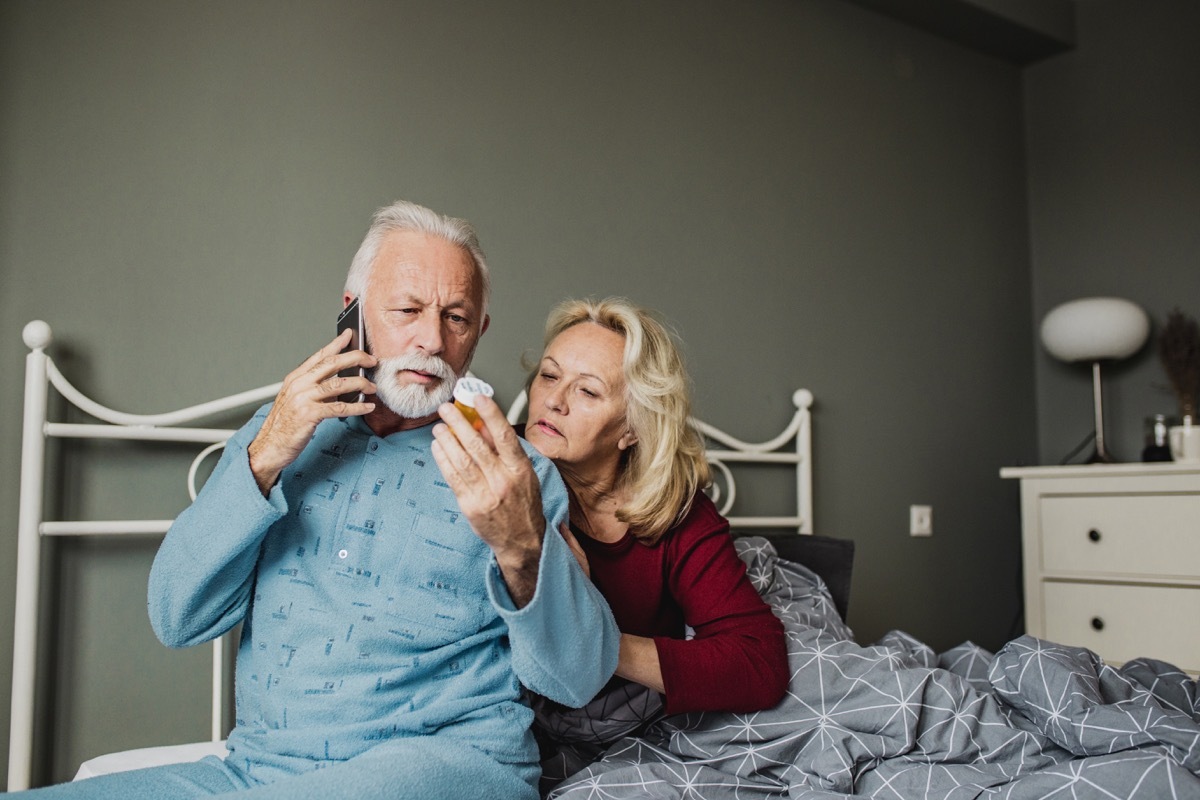 Senior couple is laying in bed, men is holding a medicine and calling a doctor