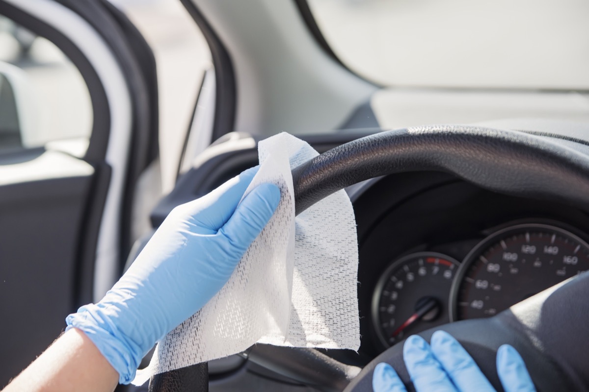 Female hands with blue glove wiping car steering wheel with disinfectant wipe. Horizontal outdoors close-up with copy space.