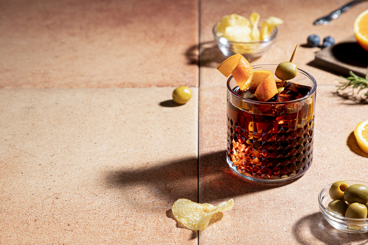 A closeup of a vermouth cocktail with an orange zest and green olive garnish, surrounded by potato chips