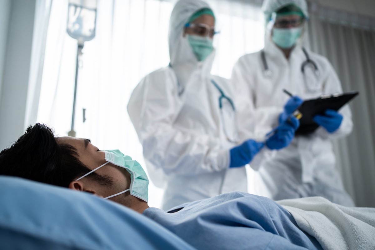 Doctor in protective suit diagnosing COVID patient on bed in hospital