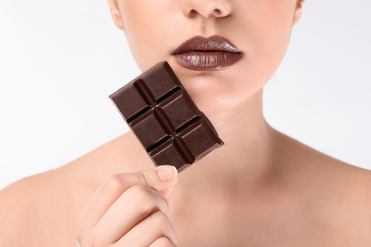 Close up of a woman wearing brown lipstick and holding a chocolate bar up to her lips