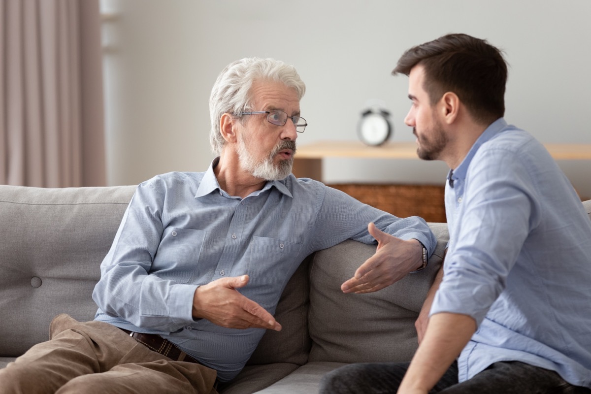 Serious 60s elderly father and grown up adult son sitting on sofa talking having important conversation trying to solve life issues problem, different men relative people communication at home concept