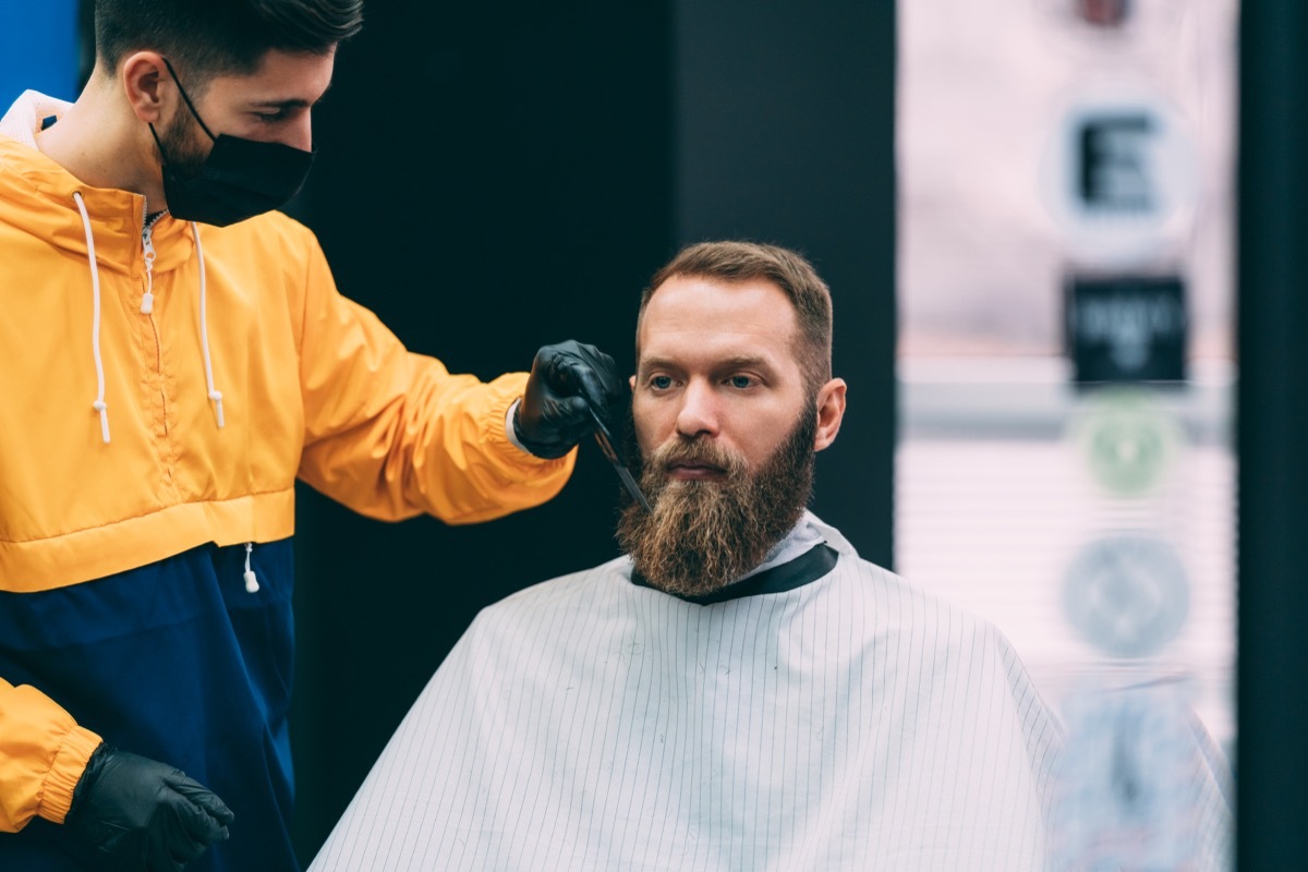 Barber with face mask combing customer‚Äôs beard