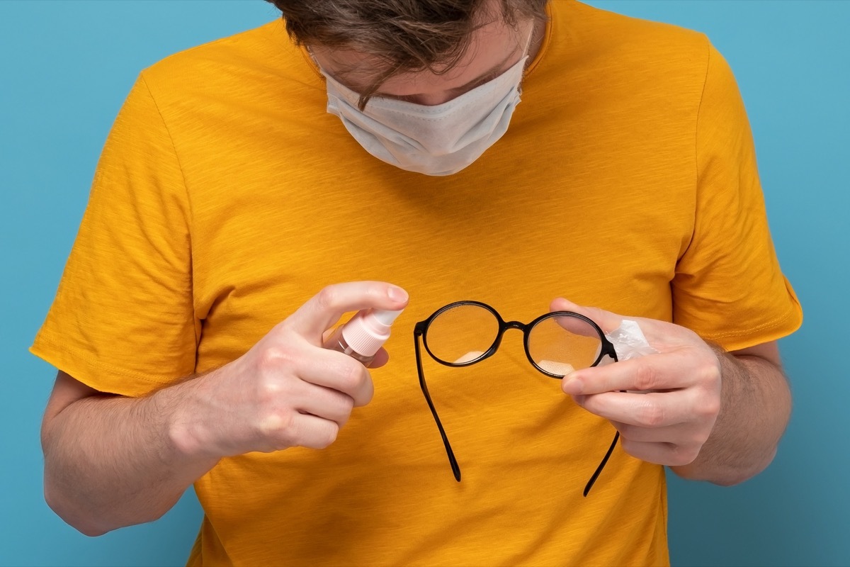 Caucasian man in medical mask disinfecting the glasses. Precaution against coronavirus or other infection. Studio shot on blue wall.