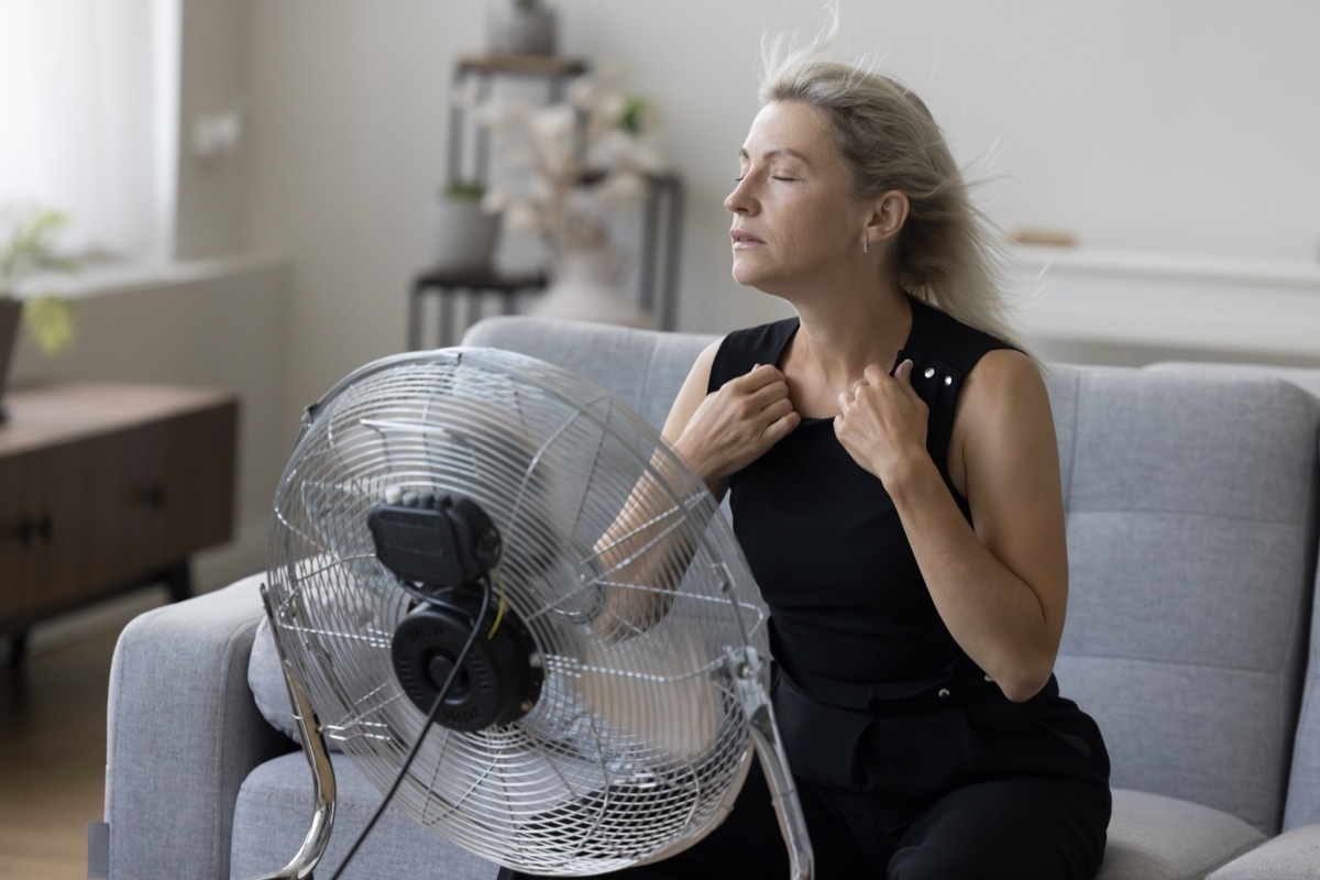 Sick exhausted senior woman with flying blonde hair and closed eyes hair sitting on couch at fresh air blowing from propeller, getting cool, refreshed at fan, feeling overheated, stress relief