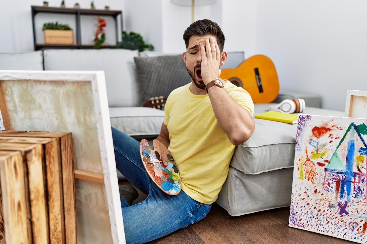 Young man with beard painting canvas at home yawning tired covering half face, eye and mouth with hand.