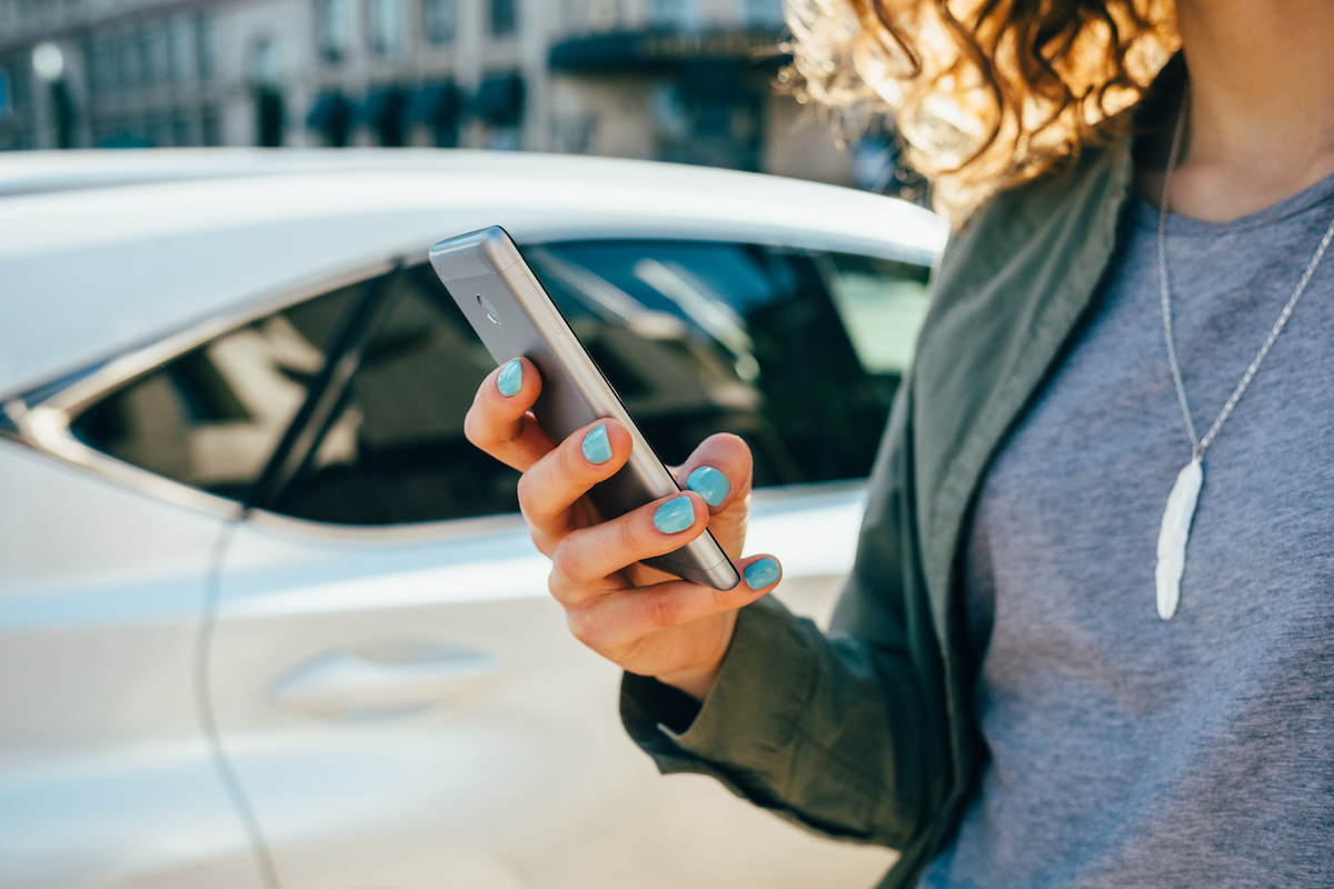 Woman using smart phone standing on street near road and car.