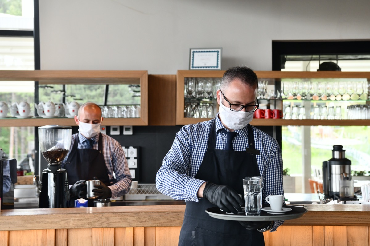 waiter in a medical protective mask serves the coffee in restaurant durin coronavirus pandemic representing new normal concept