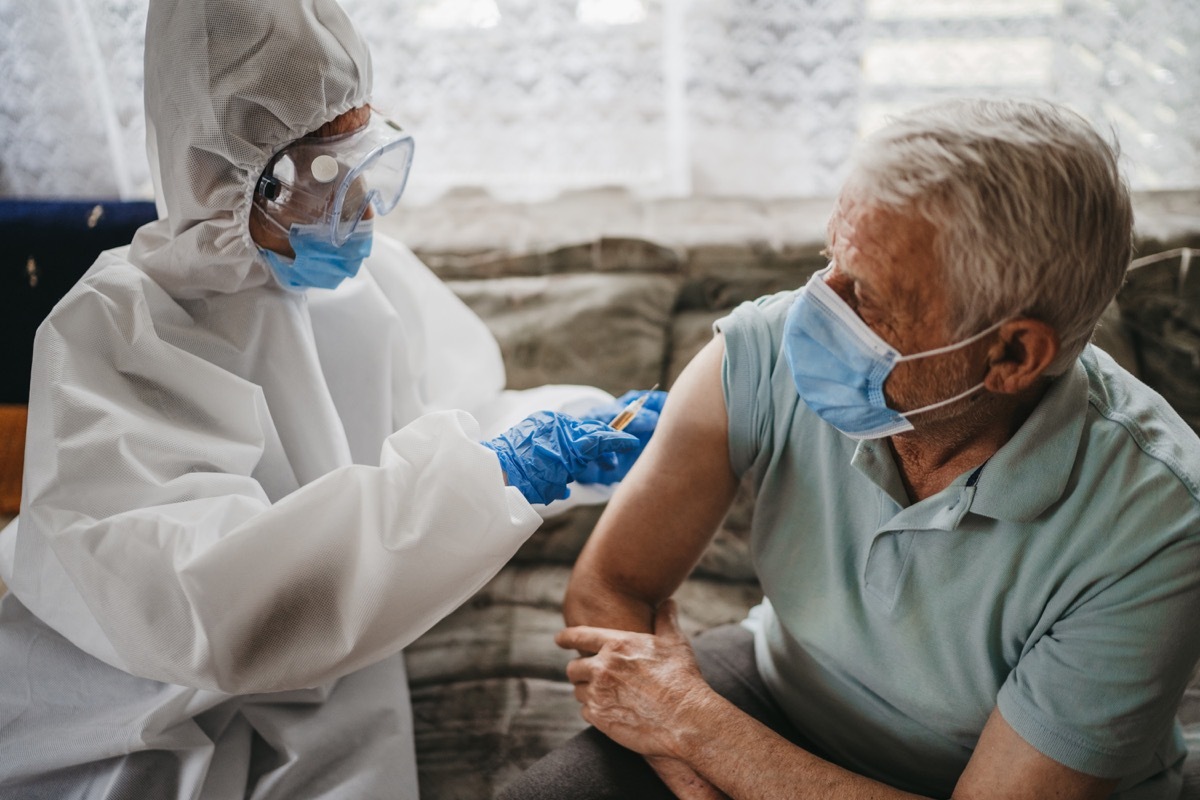 Female doctor in protective suit giving vaccine against covid19 virus to senior man at home