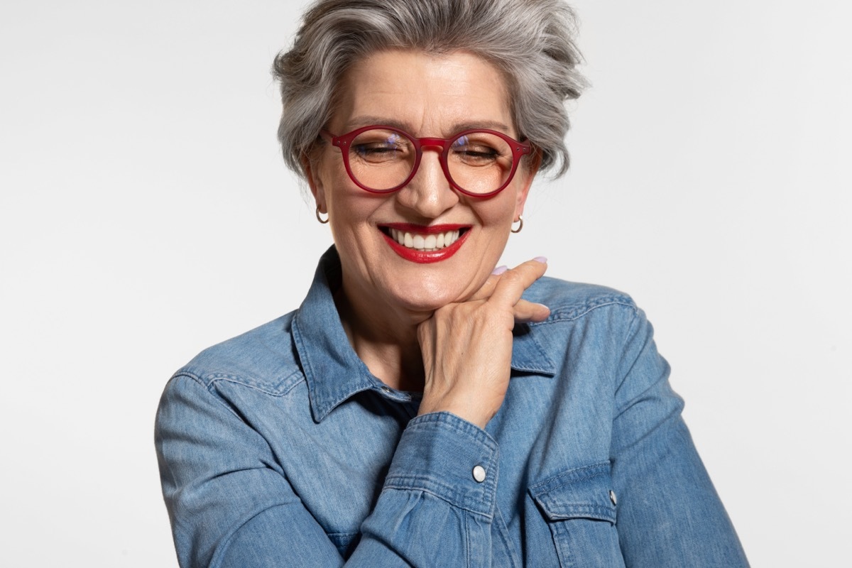 Happy Older Woman with Gray Hair wearing a denim shirt and red glasses against a light gray background