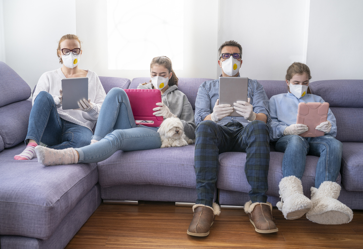 A mother, father, and their two daughters sit on a couch using tablets and devices while wearing face masks.