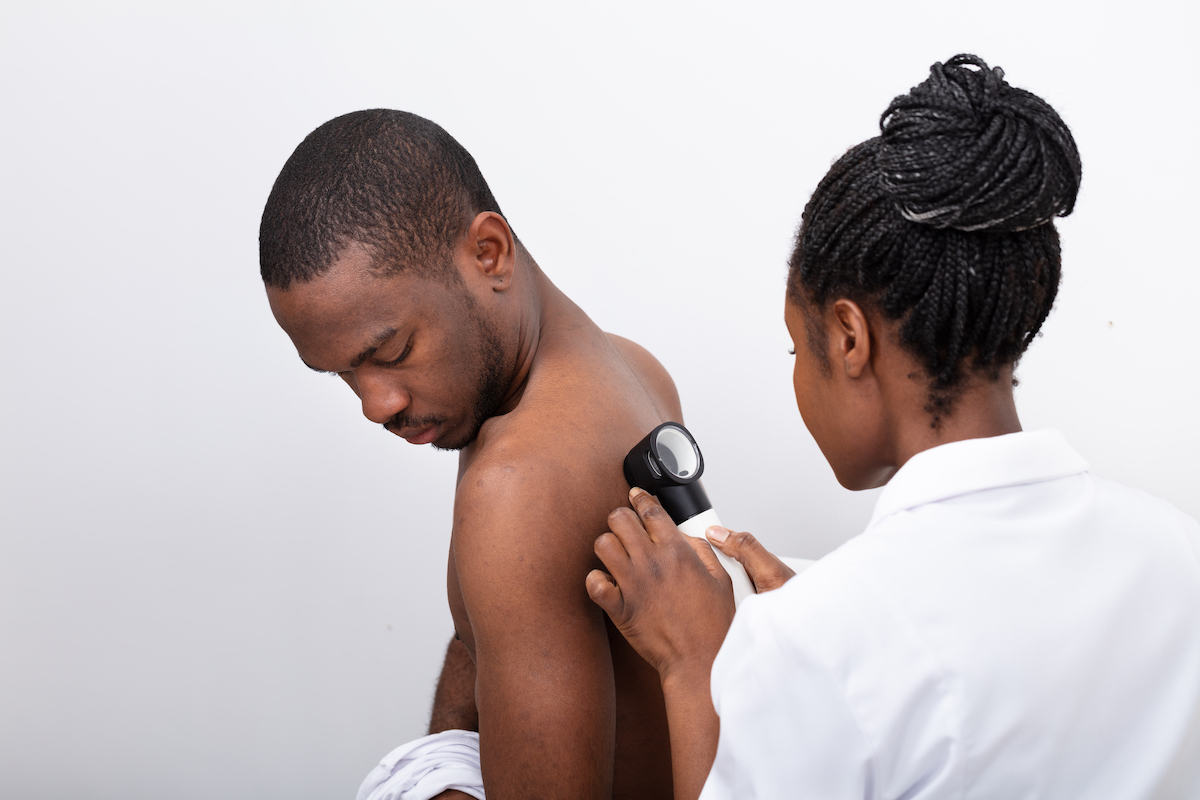 Young Doctor Examining Mole On Man's Back With Dermatoscope