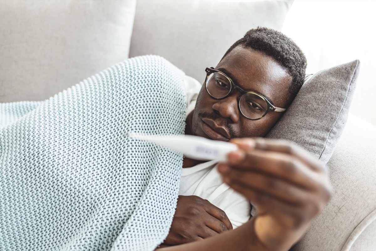 Shot of a young man checking his temperature while lying on the couch at home