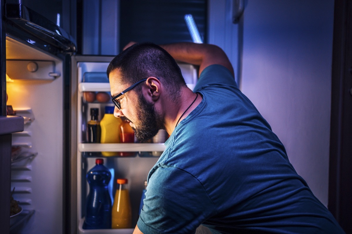 Young cheerful woman standing in front of the refrigerator at night, thinking about what to eat