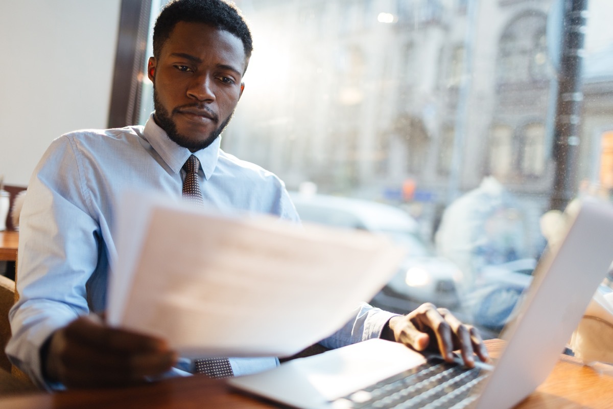 man looking over someone's resume while sitting at his laptop next to a city window