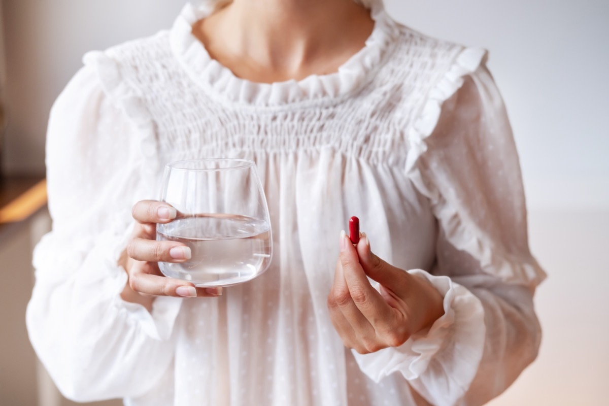 Woman in white blouse holding in hand iron Ferrum supplement capsule and glass of water. Bioactive additive woman pharmacy. Vitamin mineral treatment against anemia. Autumn health care concept