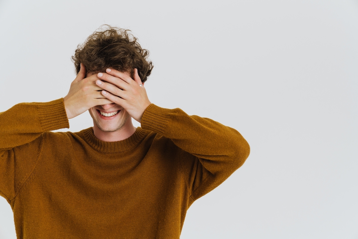 A young man with curly brown hair wears a brown sweater and covers his eyes