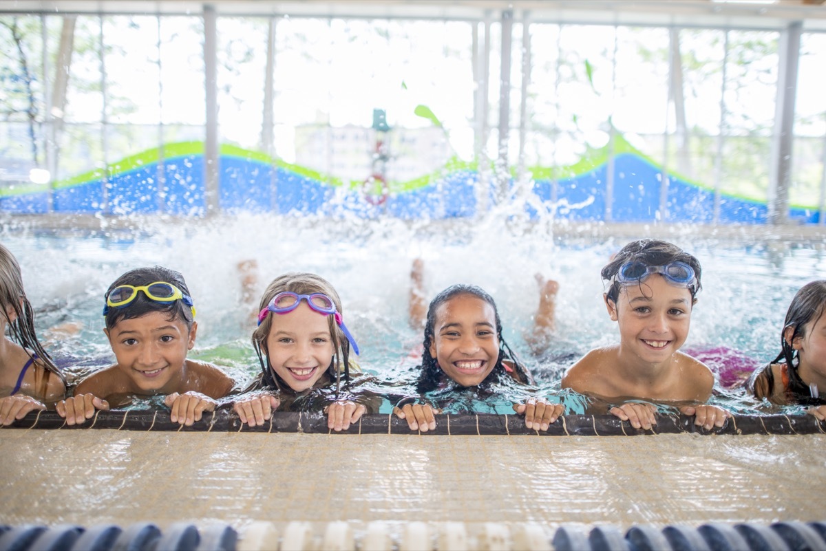 group of kids are taking a swimming class at an indoor pool. They are practicing kicking at the side of the pool, while smiling at the camera.