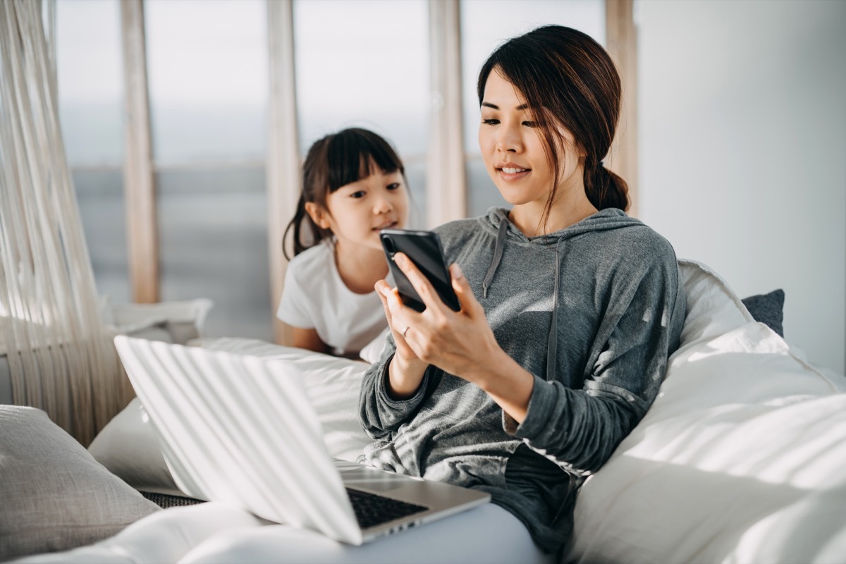 mother sitting in bed using smartphone and working on laptop