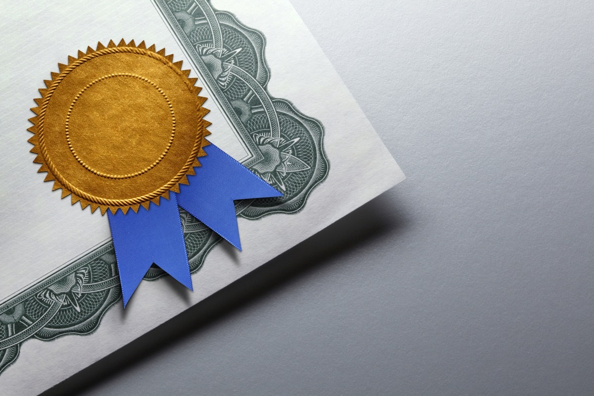 A close up of a gold seal adorned with a blue ribbon is attatched to the corner of a certificate of achievement which rests on top of a gray background which provides ample room for copy or text.