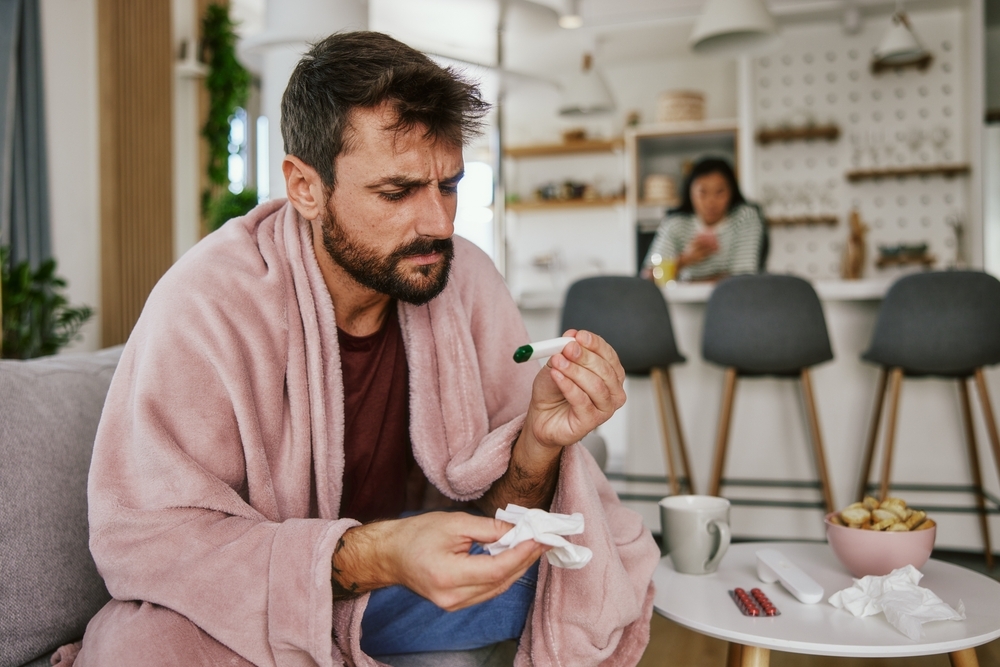 A young man sitting on the couch checking a thermometer who is sick, maybe with COVID symptoms