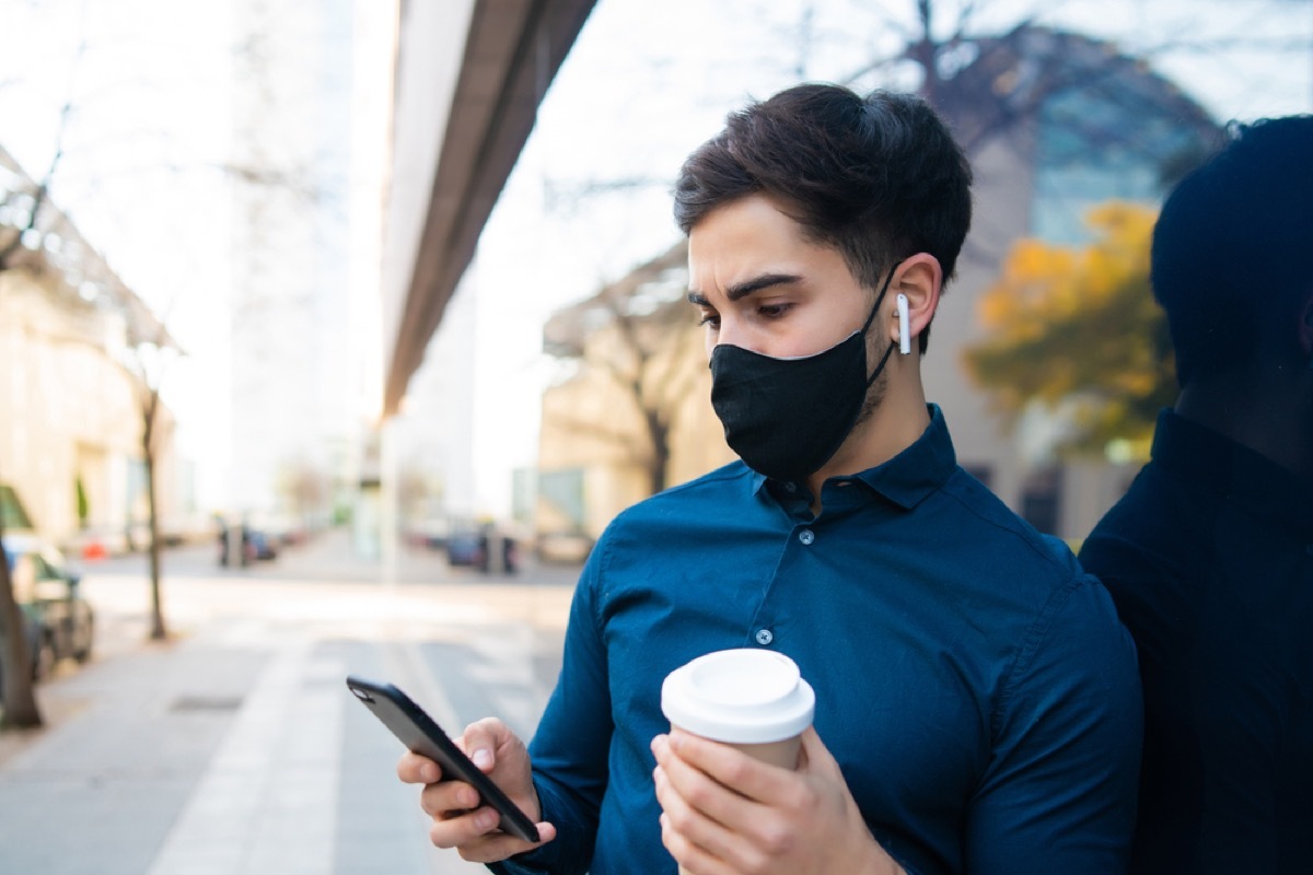 man with face mask and coffee looking at his phone outside